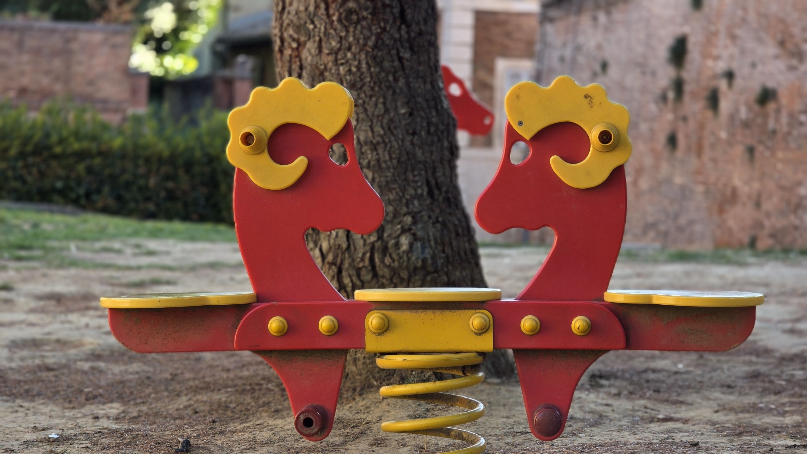 Two red rocking horses in a playground beside an old brick wall
